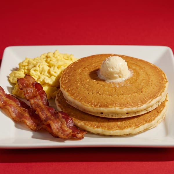 Breakfast plate with a stack of pancakes topped with butter, scrambled eggs, and crispy bacon.